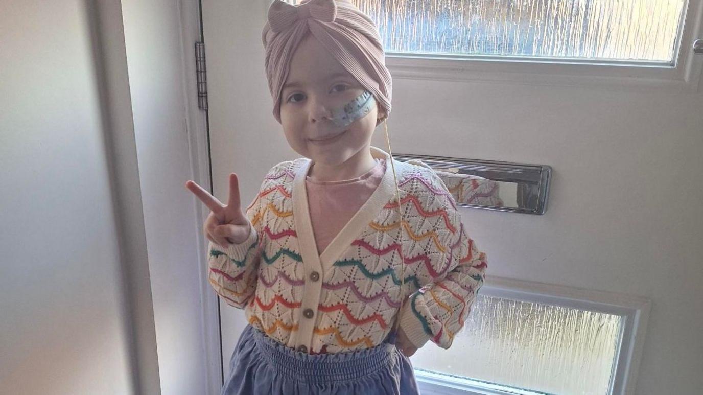 Nina is wearing a pink headscarf with a bow and a colourful jumper and is posing for the camera, inside a house and by a front door, with a victory sign.