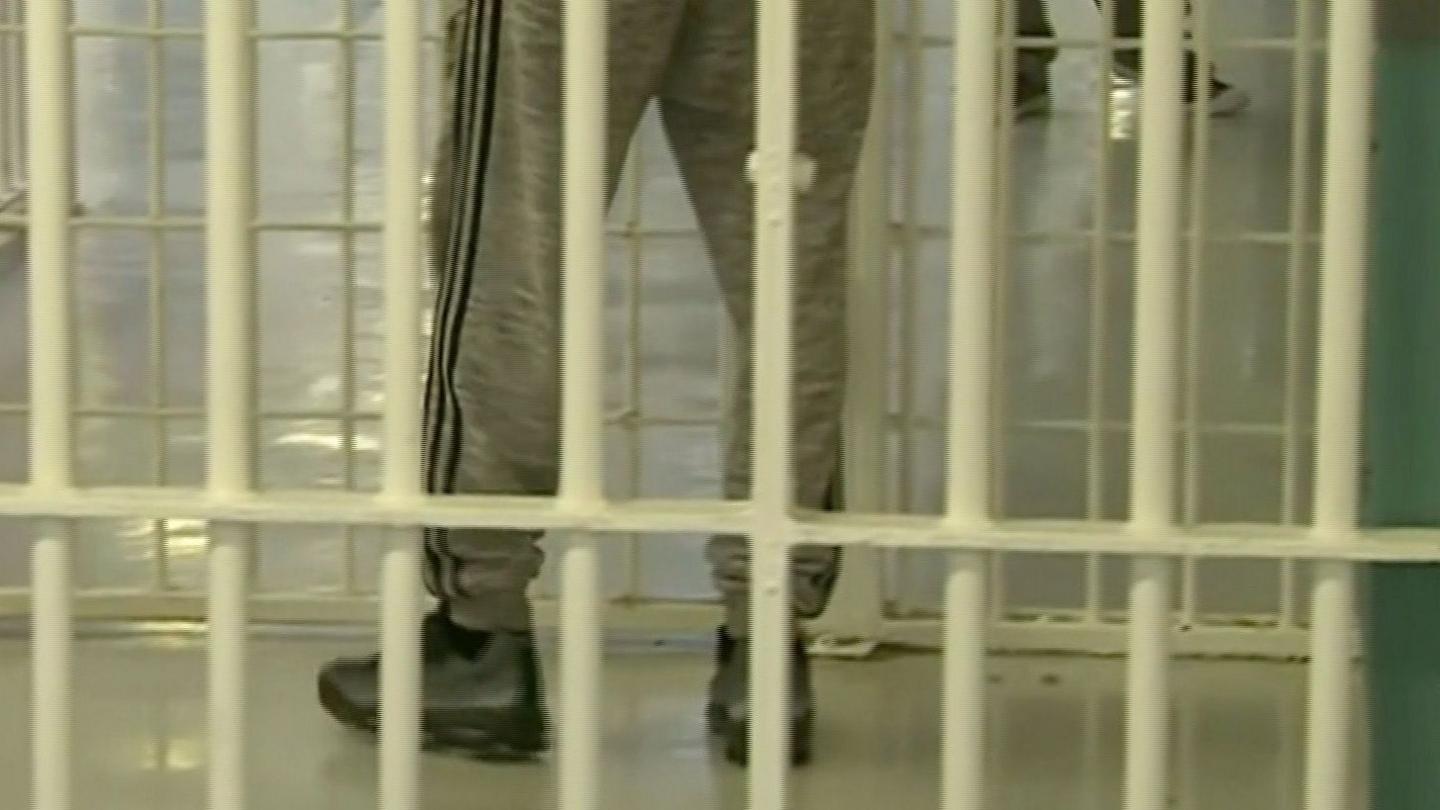 An image shows yellow painted prison bars with a person standing behind them with only their legs visible. They are wearing light grey tracksuit trousers with a double stripe down the side of the legs in a darker colour. They are also wearing grey and black trainers.