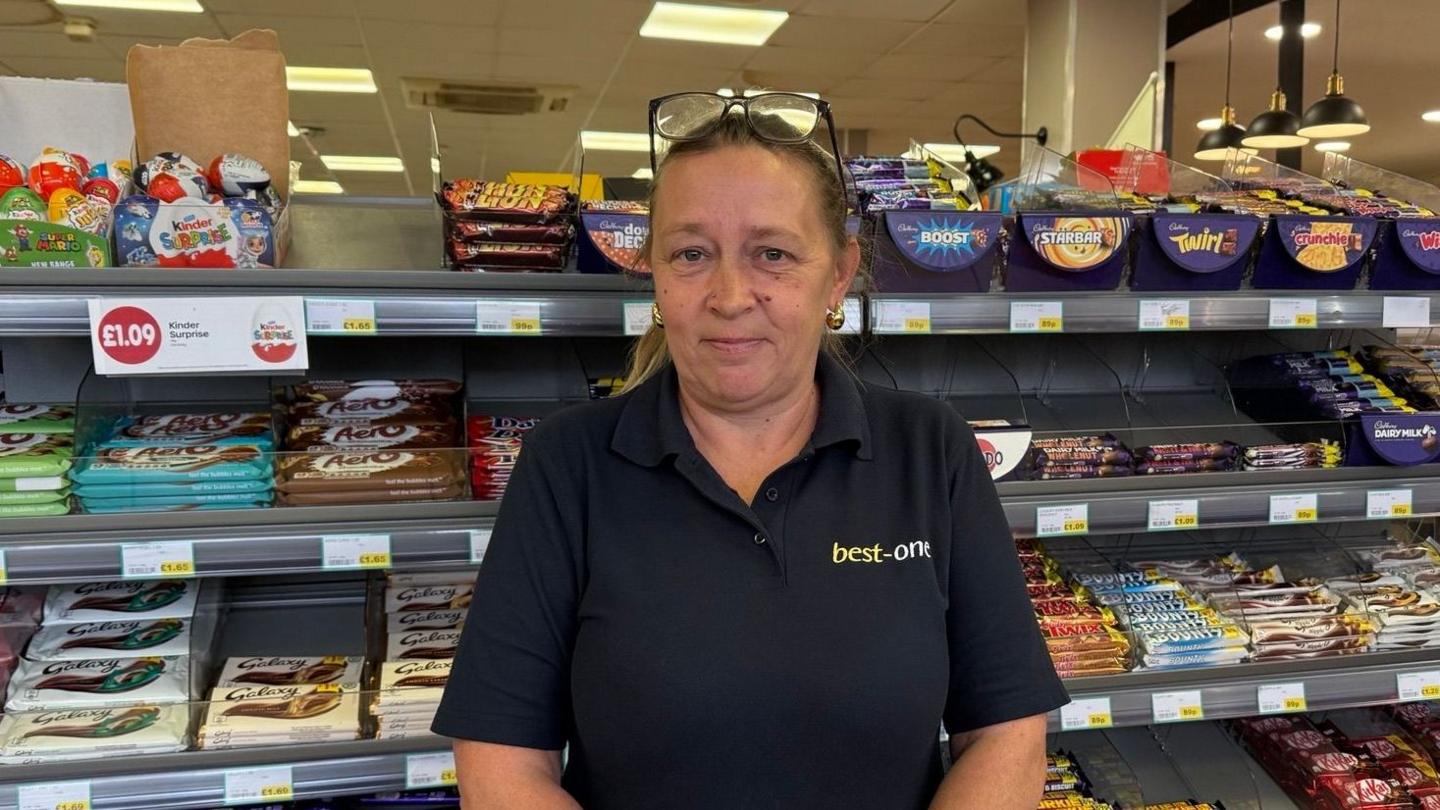 Donna has her hair tied in a ponytail and has reading glasses on top of her head. She wears a black polo shirt with 'best-one' embroidered on it and stands in front of the chocolate stand.