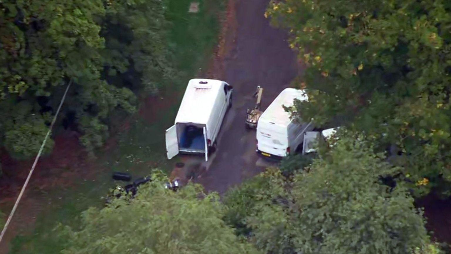 Aerial image of a remote-controlled bomb disposal robot near white vans on a country road surrounded by trees.