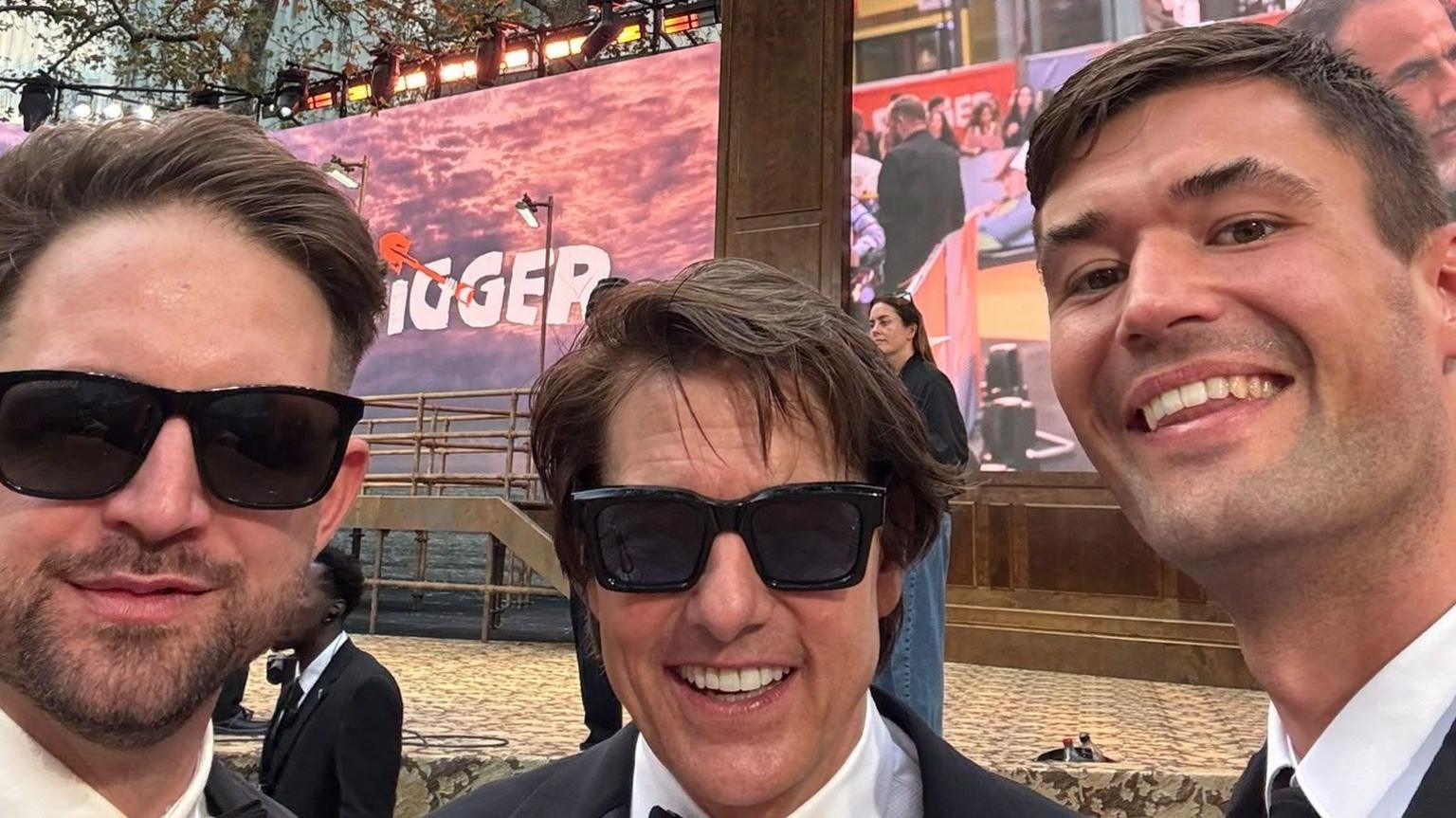 Two men dressed formally pose for a photo with actor Tom Cruise who wears a tuxedo and black sunglasses, at a film preview