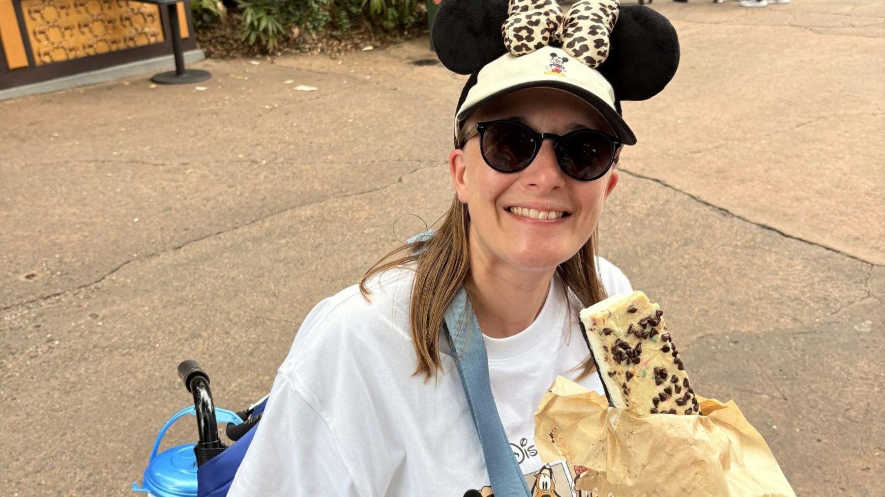 Jennifer sits in a wheelchair. She is eating a sweet treat from a paper bag and is wearing a white Disney branded T-shirt and a cap with Mickey Mouse ears. She has sunglasses on, her long hair falls below her shoulders, and she is smiling.