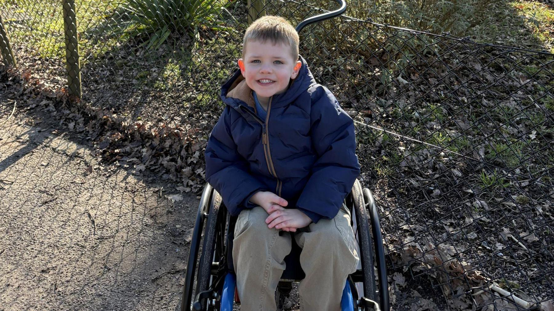 William sitting in a wheelchair. He is wearing a dark navy coat and khaki trousers and is smiling at the camera. There is a fence and grass behind him.