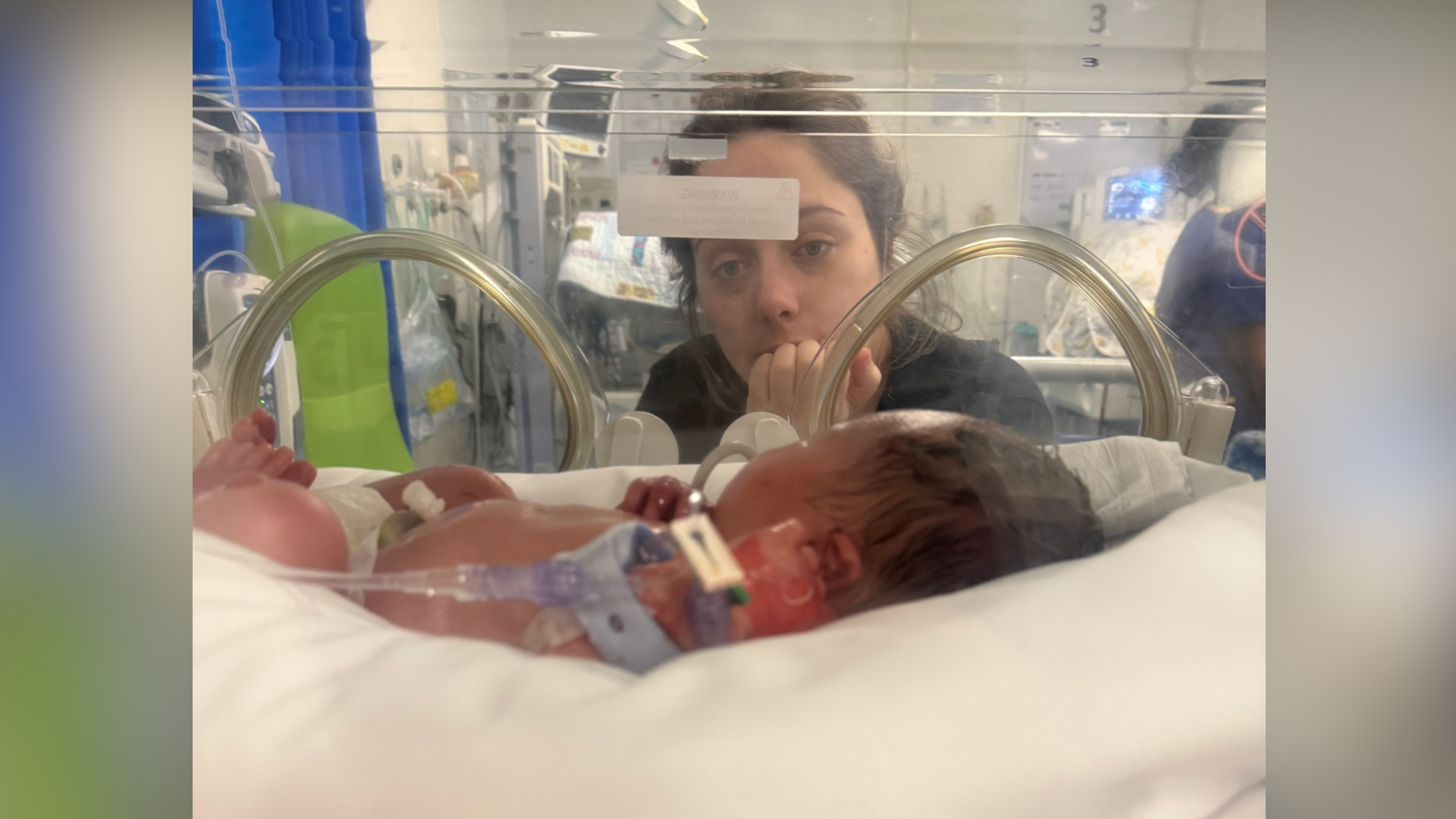 In the foreground of the picture is baby Ralph who is inside an incubator with wires hooked up to his body. Behind the incubator Ciara, his mum who has brunette hair and is wearing a black T-shirt, is looking into the incubator at her son.