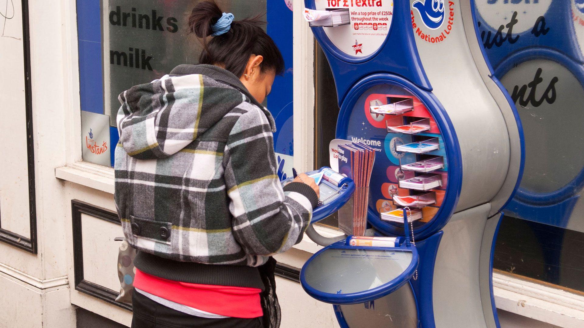 A woman fills out a form for a National Lottery ticket at a stand outside a shop