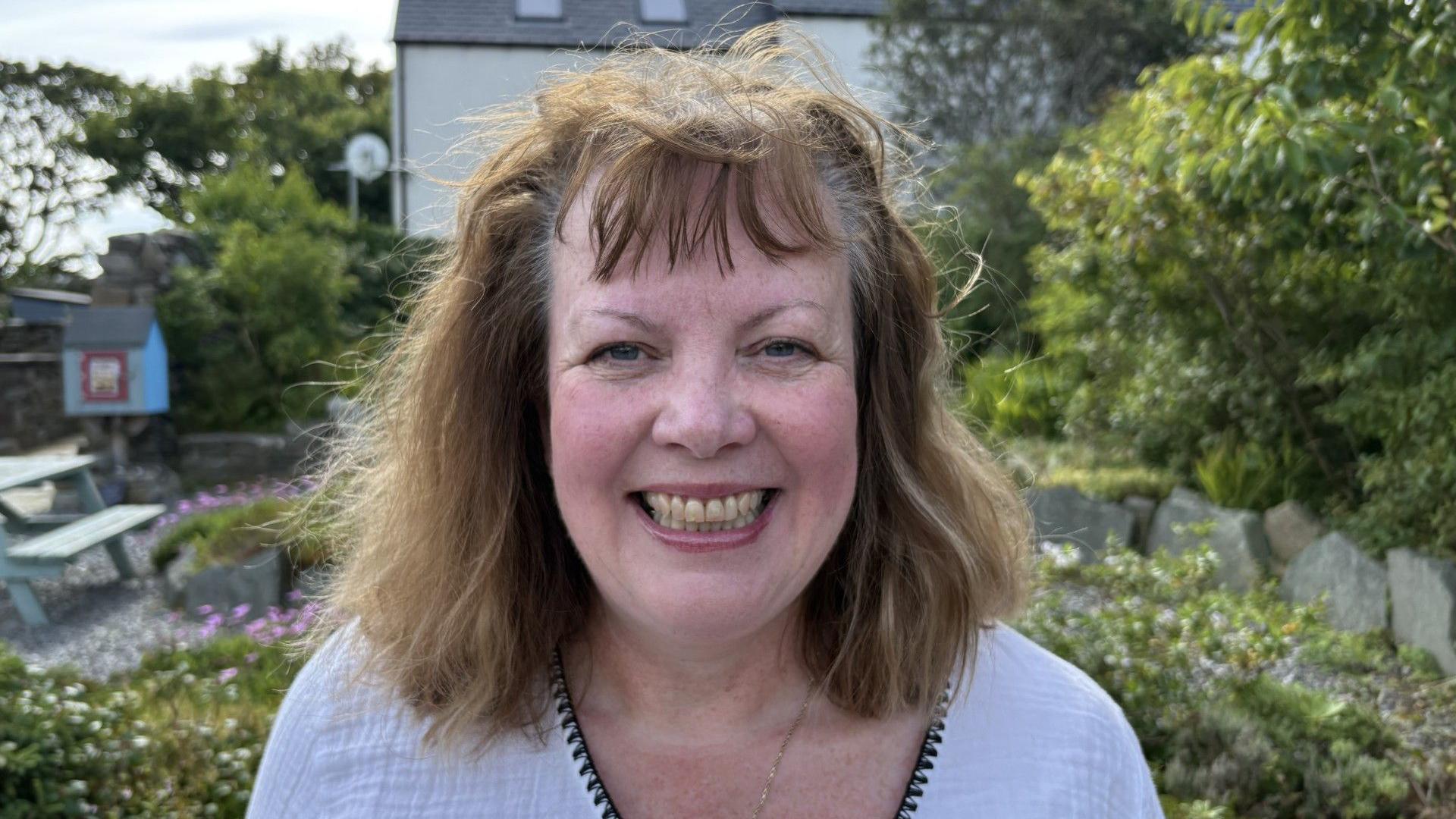Lorraine Hannah is a middle-aged woman. She is smiling at the camera and standing in a public garden. She has brown hair and is wearing a white top. There's a picnic bench in the background and it's a sunny day.