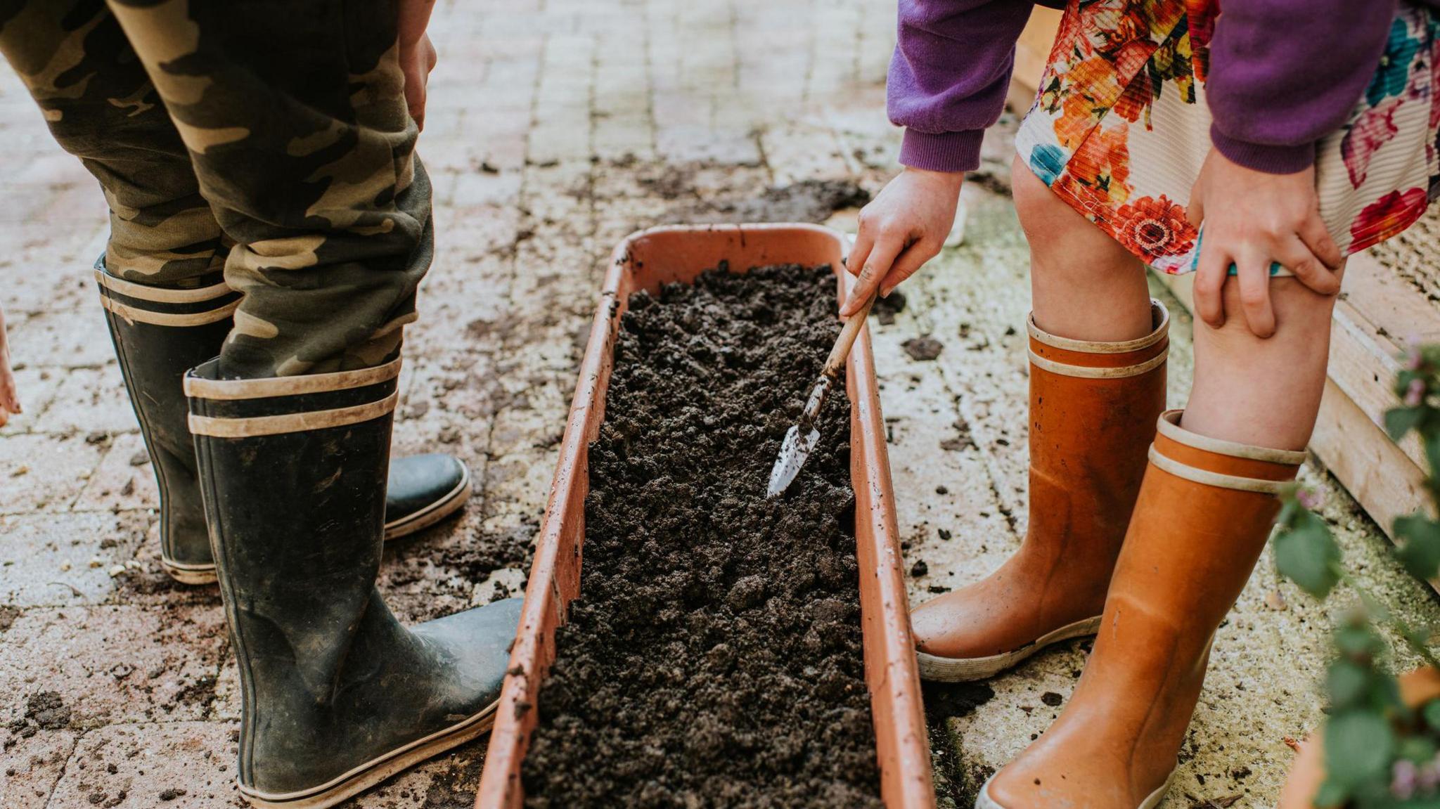 Focus is on the wellies of two children - a girl and a boy, using a small trowel to plant seeds in a long planter trough.
