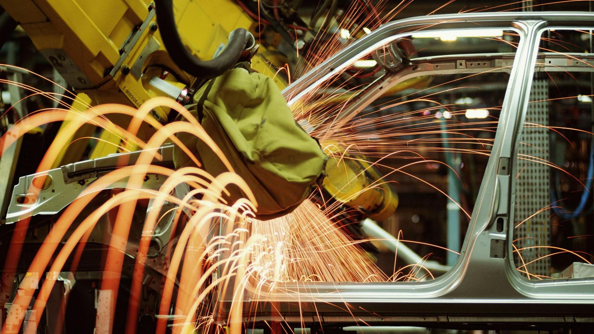 A car being manufactured. A yellow robotic arm is working on the car chassis, generating dozens of orange sparks.