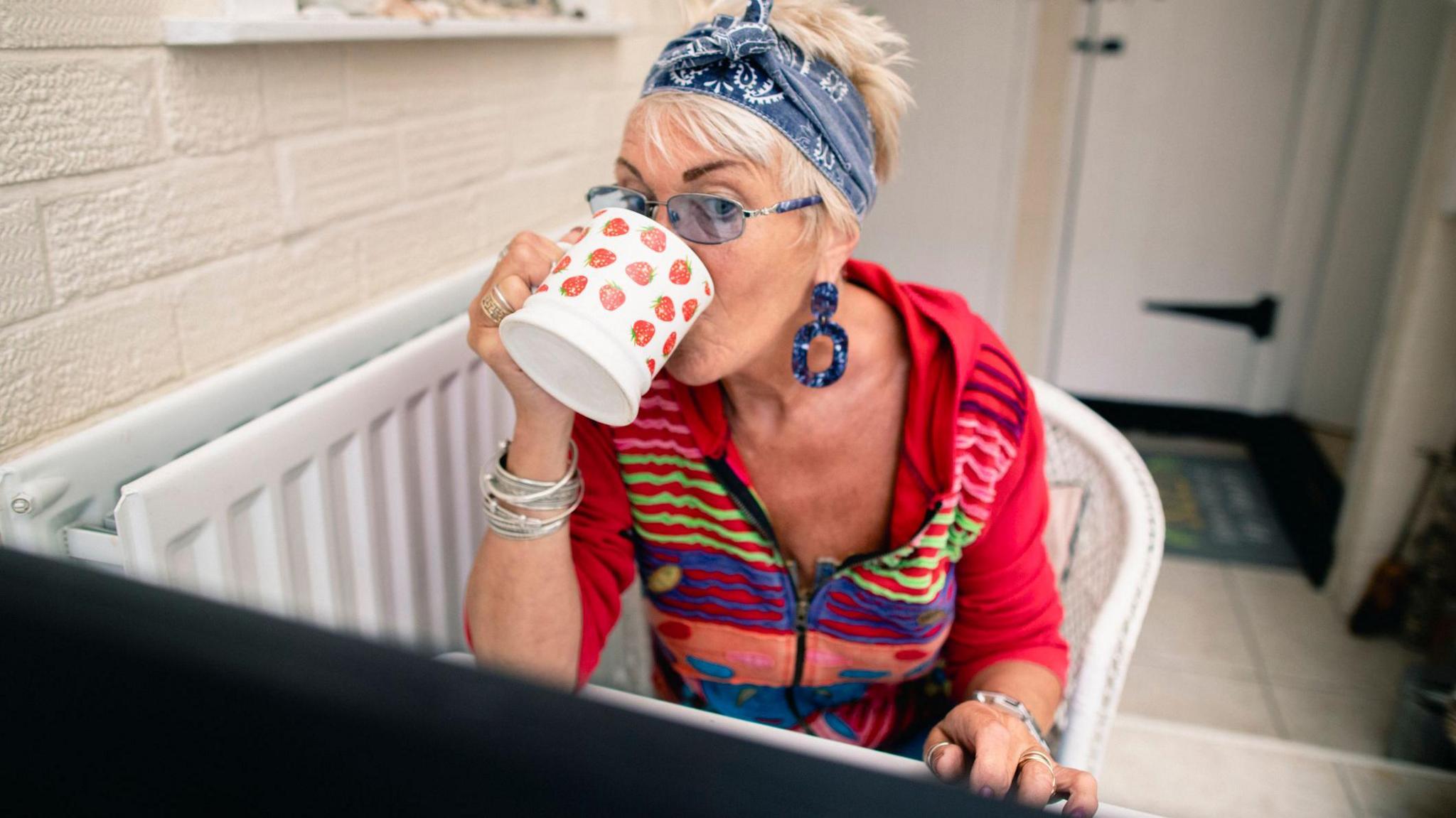 Woman wearing colourful cardigan and drinking from a mug sits behind a computer screen.