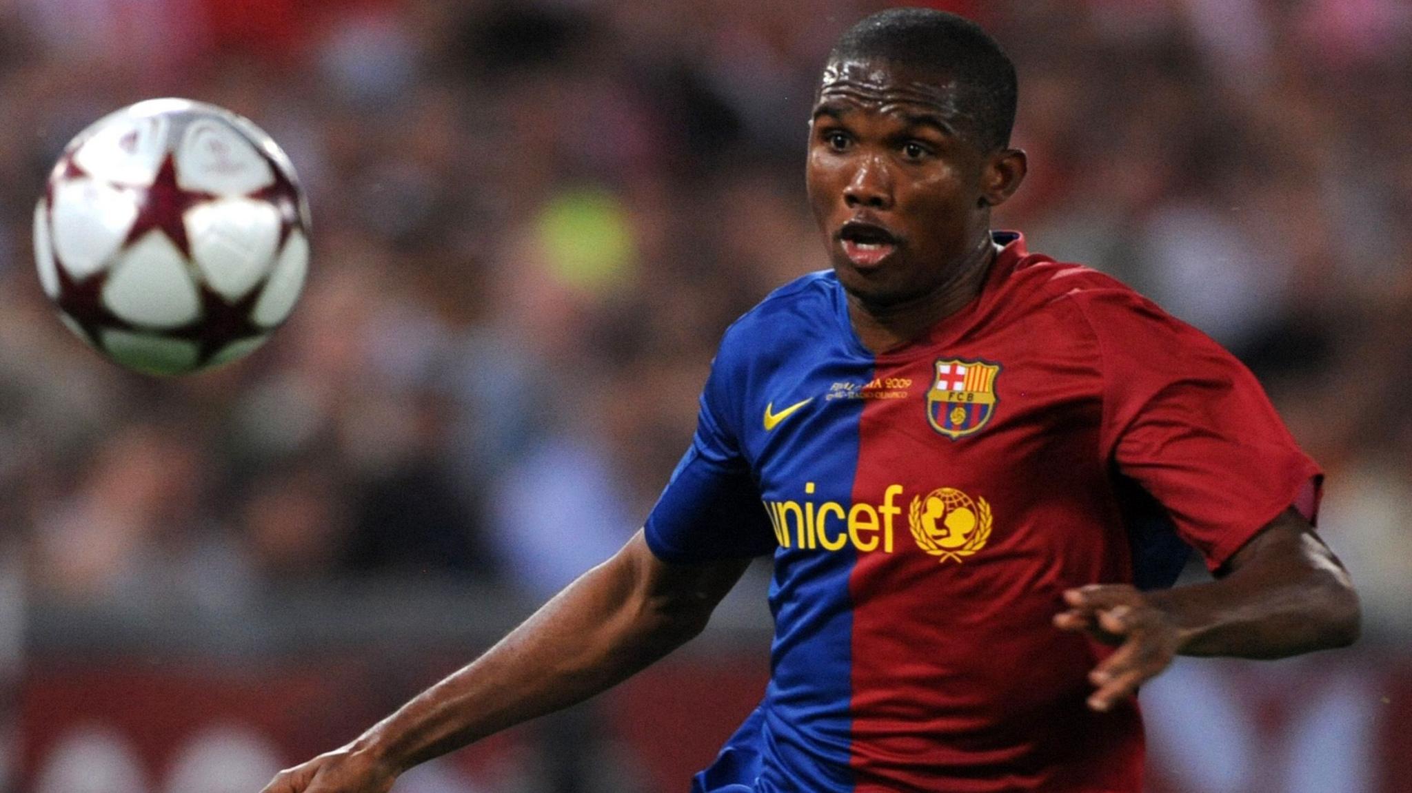 Samuel Eto'o, visible from waist up wearing a blue and claret Barcelona home shirt, chases after a Champions League-branded football which is hanging in the air. His gaze is firmly locked in on the ball
