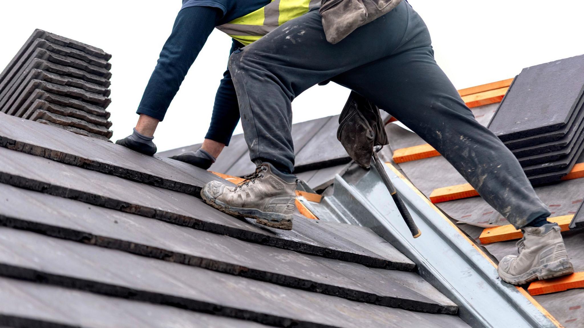 Lower half of a workman standing on a roof with a chimney visible. Some tiles are not yet laid on the roof structure.
