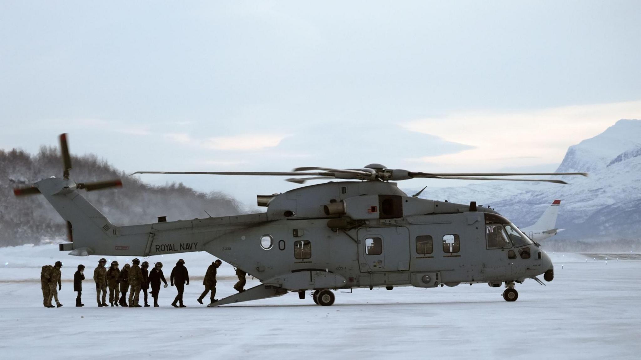A group of people board a grey army helicopter in Norway with snowy mountains in the background