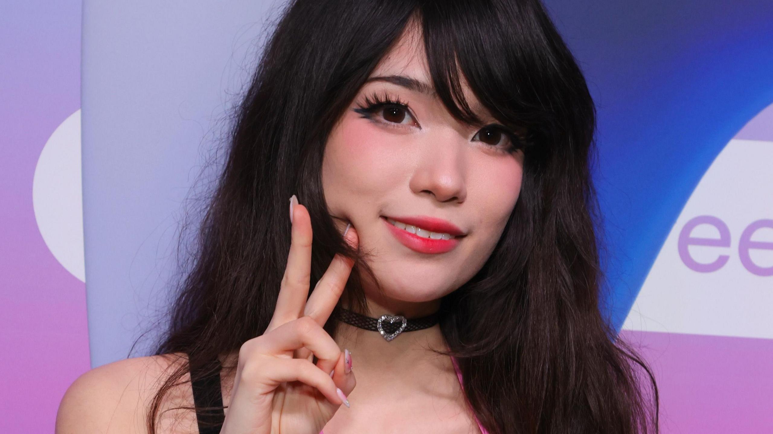 Emiru at TwitchCon 2024. She has long dark hair worn loose and wears false eyelashes, red lipstick and blush. She makes a peace sign while smiling at the camera.