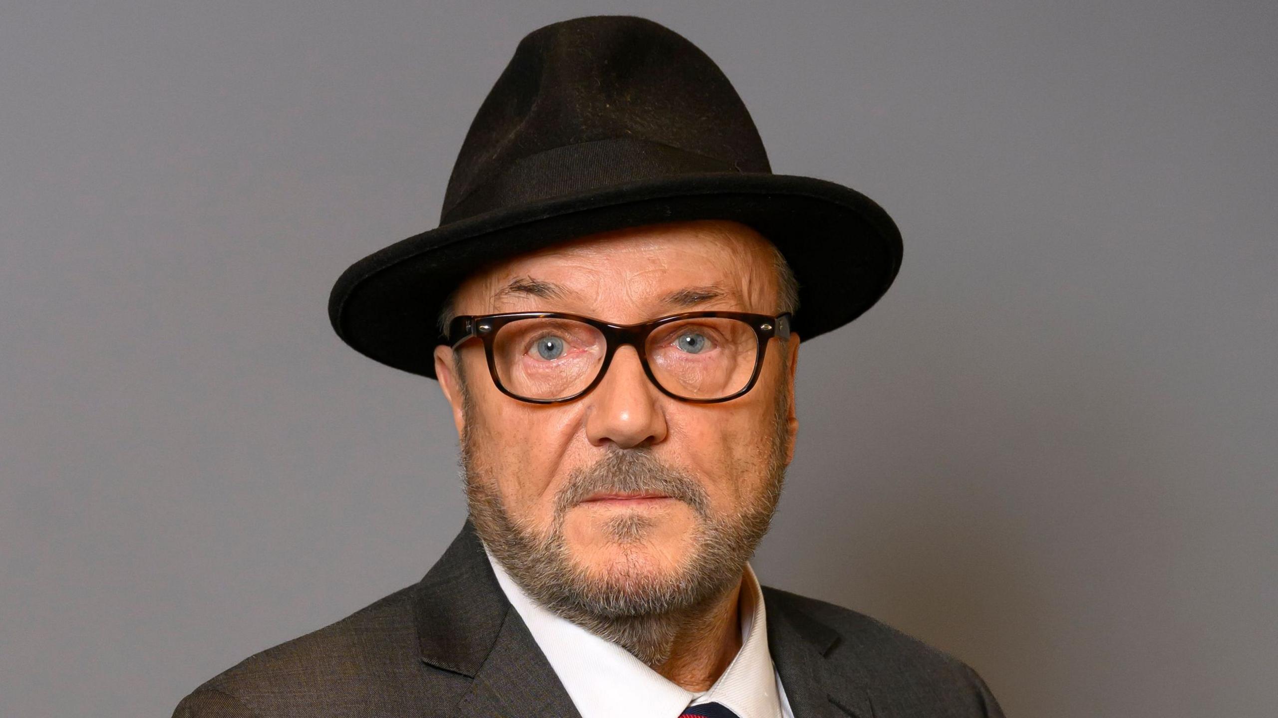 Official portrait of George Galloway wearing a dark suit, white shirt, patterned tie, black fedora hat, and dark-rimmed glasses, set against a plain grey background.