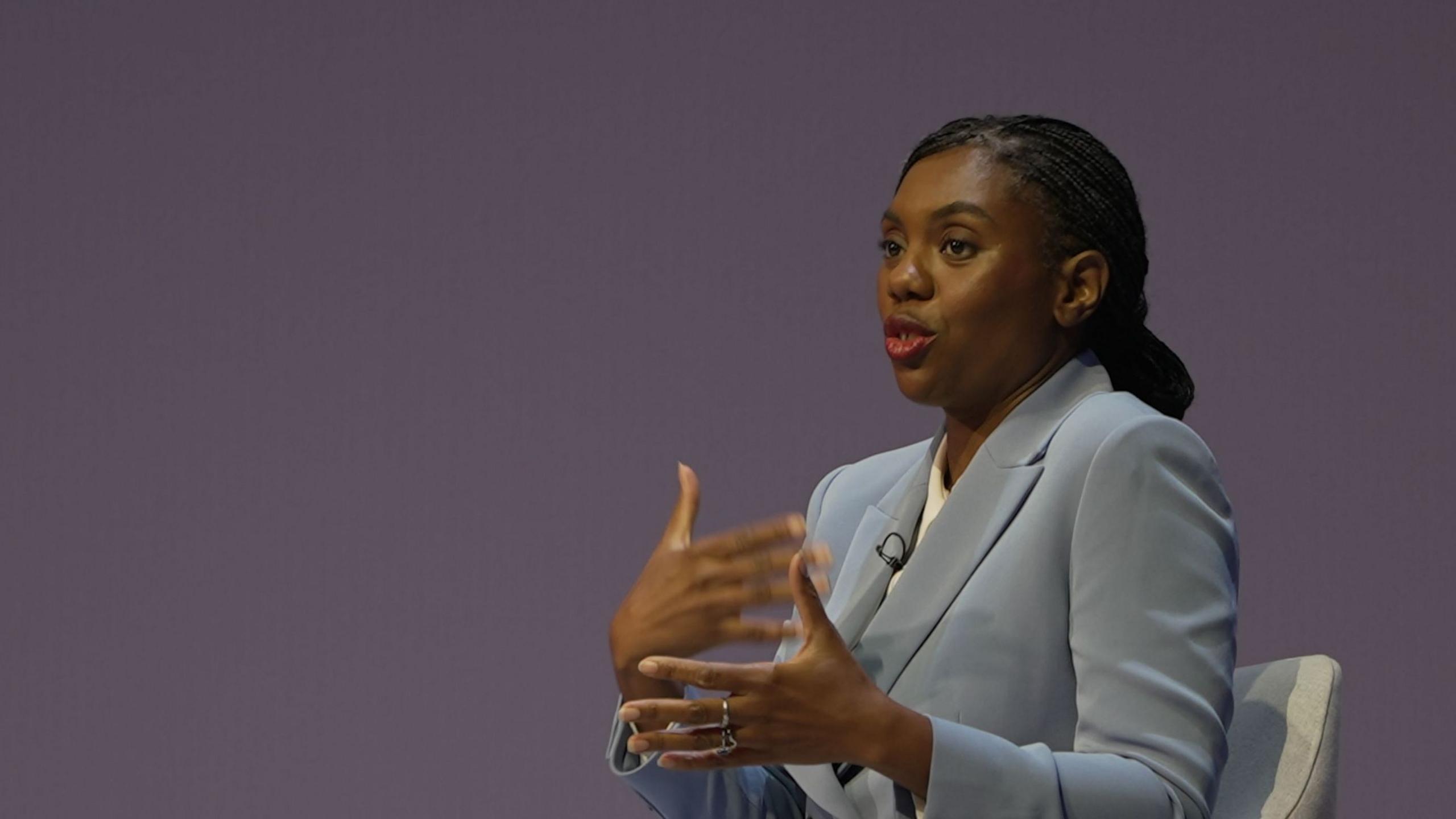 Kemi Badenoch on stage at the Conservative Pary conference, speaking and gesturing with her hands. She is wearing a pale blue suit, with a white shirt and her hair is tied back.