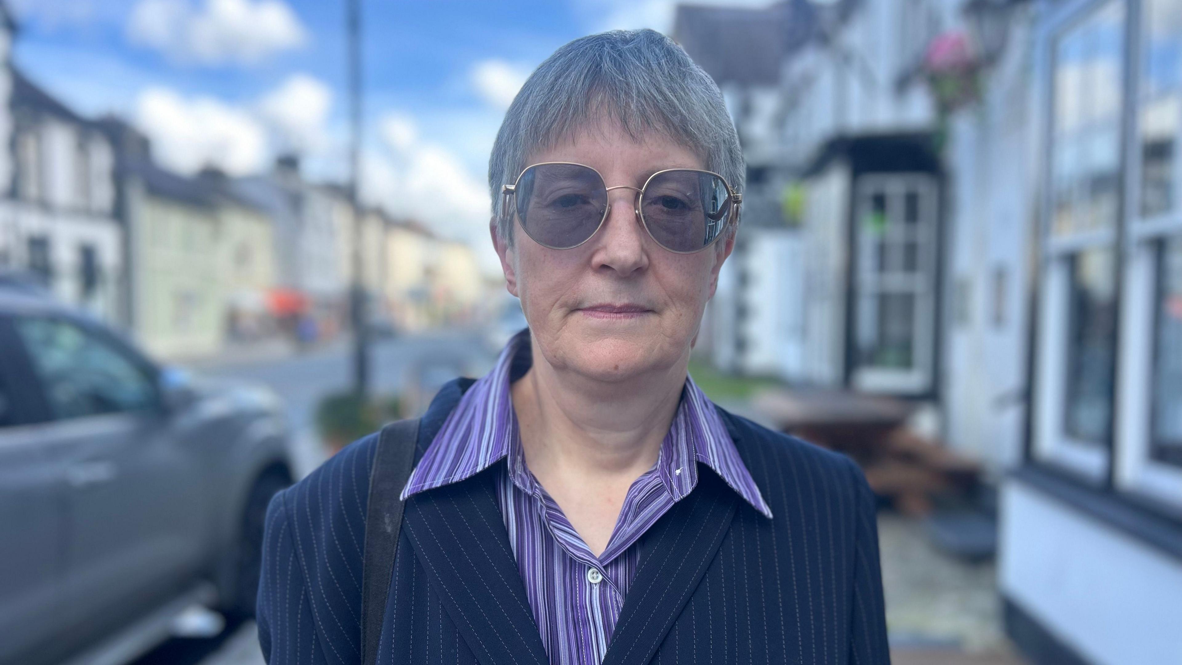 Kate Baxter with dark glasses and short grey hair. She's wearing a purple striped shirt and a navy striped blazer. She has a handbag strap on her shoulder. She's standing on a high street.