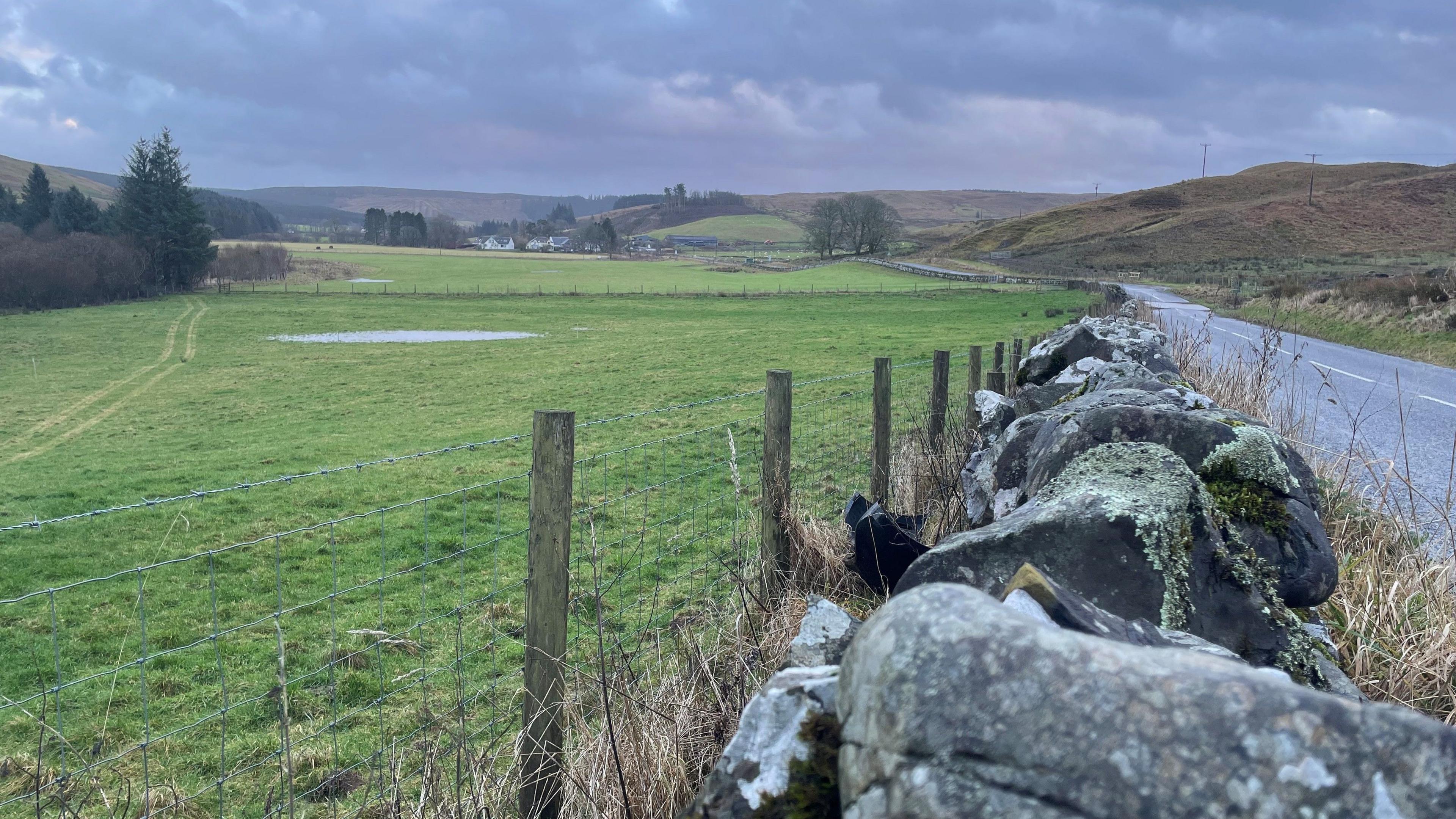A view over a dry stone wall and fence across fields. The village of Eskdalemuir is in the distance. In the corner of the image is a narrow winding road