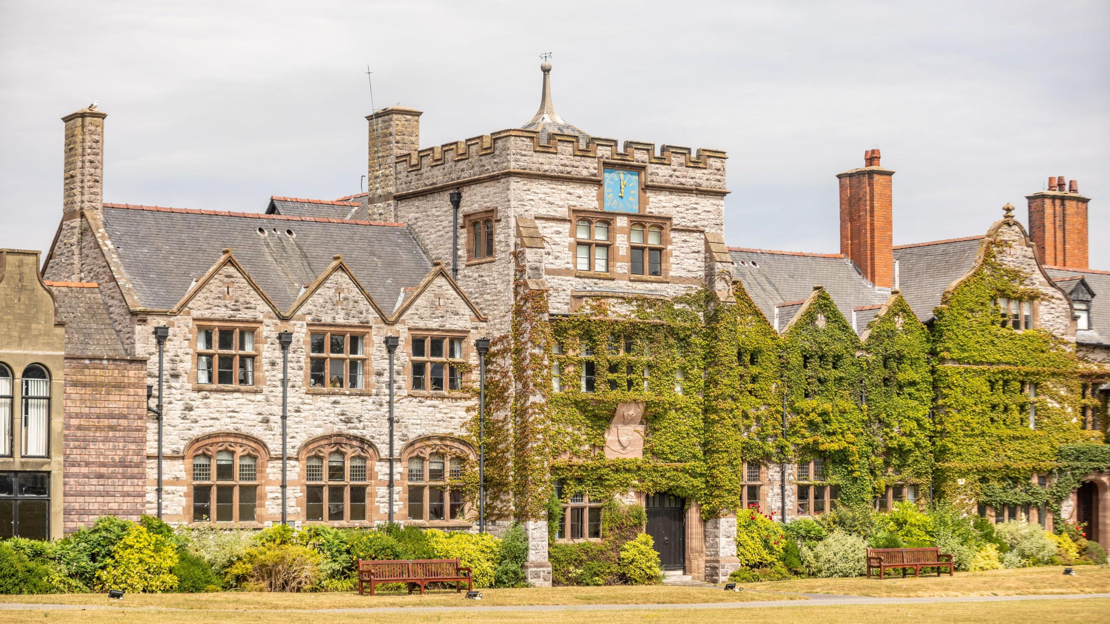 A historic school building constructed from pale stone stands behind a grassy lawn, with climbing ivy covering much of the frontage and chimneys rising above the roofline.