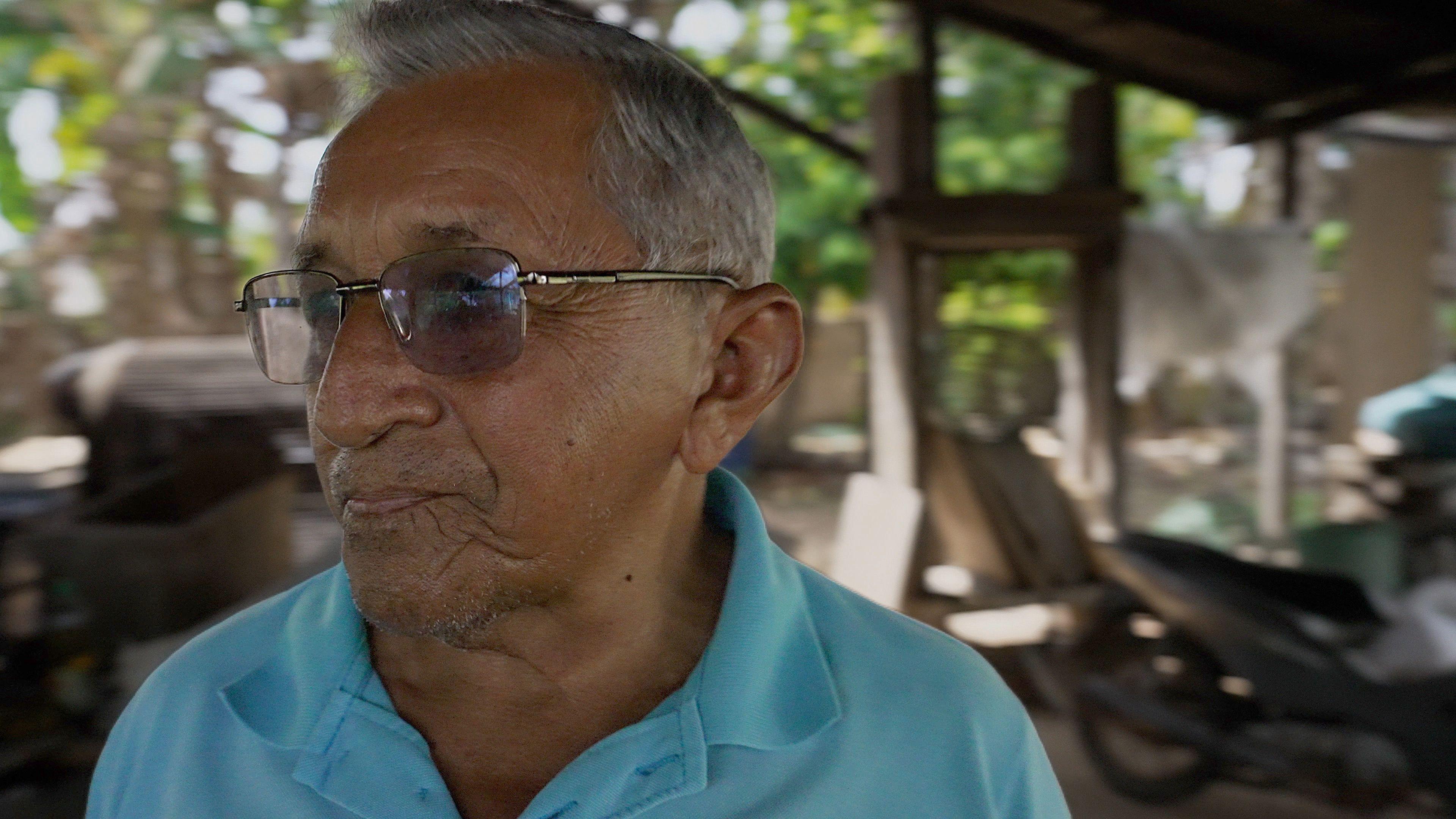 A man with siver hair and tanned skin wearing dark sunglasses and a light blue polo neck t-shirt stands in his farmyeard. In the background, you can see his wooden tools and a motorbike.