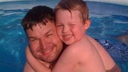 A dad and his son holding each other in a swimming pool. They are both smiling at the camera.