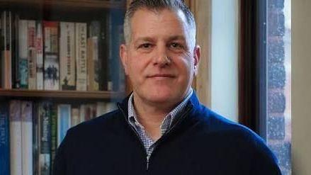 A middle aged man poses for a photo in a blue jumper and blue shirt standing in front of a bookcase.