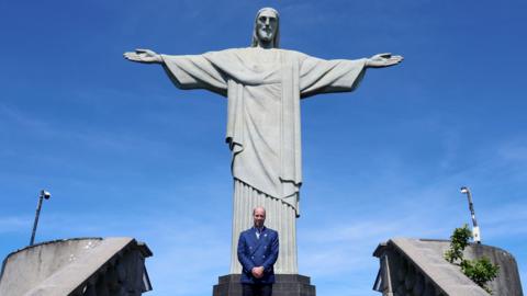 Prince William stood in front of the Christ the Redeemer statue, which looks light grey. The sky is bright blue and the prince is wearing a navy suit.