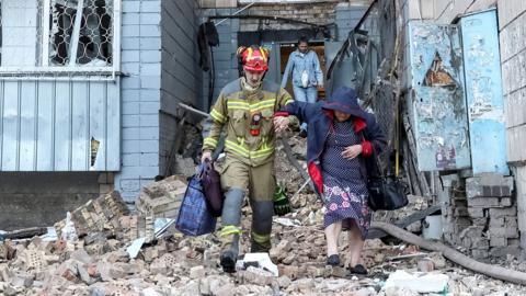 A firefighter helps an old lady out a building holding her arm as they step over piles of bricks and rubble