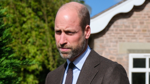 Prince William, who has a bald head and grey facial hair and is wearing a grey suit, pale blue shirt and dark tie. He is stood outside a house.