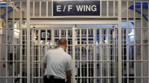 An image of a series of security gates in a prison - with floor-to-ceiling white metal bars. Above the first set it says "E/F Wing" and a prison officer - a bald man in a white shirt and black trousers - is opening the gate. We see him from behind.