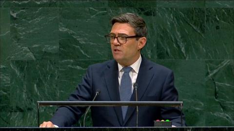 British Prime Minister Andy Burnham speaking at a lectern in a navy suit in front of a green tiled wall.