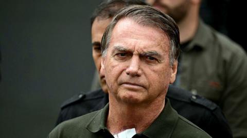 Police escort Jair Bolsonaro, who wears a green polo top, from hospital