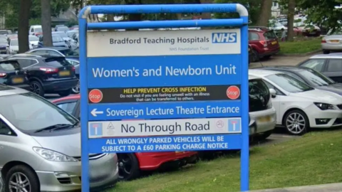 A large blue and white sign identifies the women and newborns unit at Bradford Teaching Hospitals NHS Trust. It sits in front of several rows of parked cars.