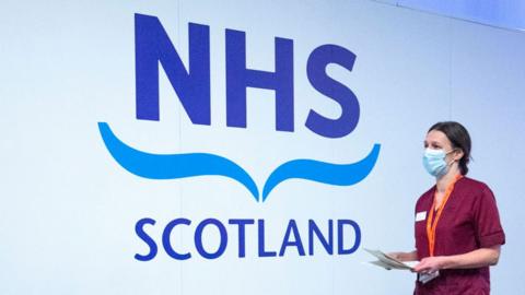 A white wall with blue lettering which says 'NHS Scotland' - a medical professional wearing maroon scrubs and a surgical mask walks by