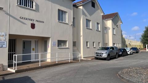 Guernsey's Les Nicolles Prison from the outside. It's a yellow building with a sign which says Guernsey Prison on it. The windows have white bars on them and in the middle is a grey/blue door.