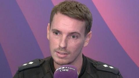 Insp Ryan Walker, a man with short brown hair and stubble, wearing his police uniform, sits in the BBC CWR studio with a BBC Radio CWR purple microphone in front of him. behind him is a purple and pink background.