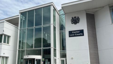 The exterior of Exeter Crown Court. The front of the building is white and has large glass windows on the left. On the right, there is a black sign with the words 'Exeter Law Courts' with a badge above.
