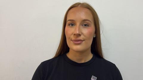 Head-and-shoulders portrait of Hannah Pritchett with long straight hair wearing a black T-shirt, standing against a plain light-coloured wall. A small badge on the shirt reads "Knife Crime". She is facing the camera directly in a well-lit indoor setting.