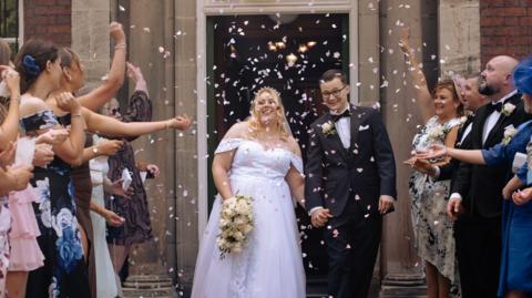 A wedding party is gathered outside a stone-fronted building as confetti is thrown into the air. Two people in formal wedding attire are walking hand in hand through a line of guests, with one carrying a bouquet of white flowers. Guests on either side are dressed in occasion wear and are tossing pink and white confetti.