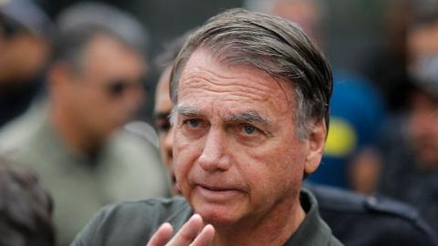 Brazil's former president Jair Bolsonaro is captured mid-wave, his fingers just visible, looking into the distance. His hair is combed over to the right and he is wearing a green khaki shirt.