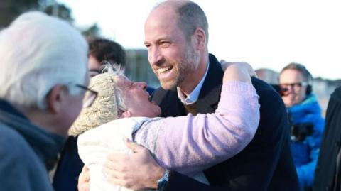 A older woman with white hair, wearing a pink jumper, gilet and a woolly hat, embraces Prince William as she looks up at him. Prince William smiles as people around them watch.