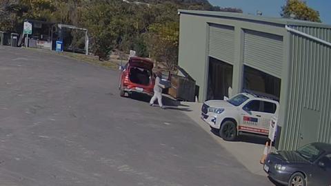 A person dressed in a grey tracksuit holding a box walks into a green shed away from red car with its boot open.