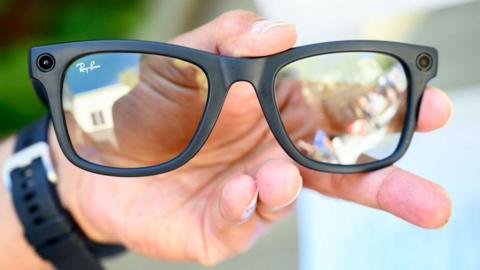A pair of Meta glasses, being held in someone's left hand. They are black with a camera visible on each side. The person is wearing a watch.