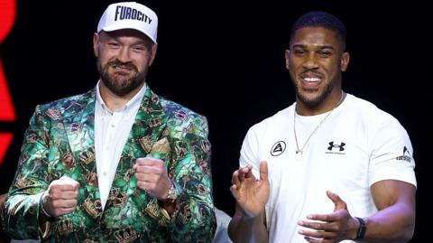 Tyson Fury holds up his fists and Anthony Joshua holds up his hands and smiles