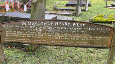 A wooden memorial stands in a graveyard and bears the inscription 'in memory of Henry West'