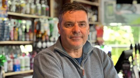 Mark, who has short dark hair and is wearing a grey jumper. He is standing in front of a bar.