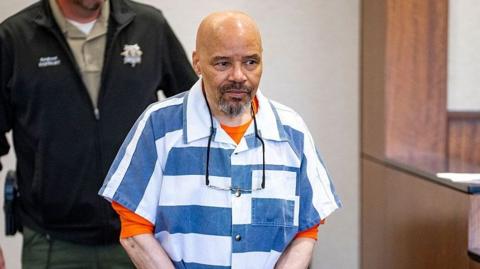 Douglas Carter appears for a bond hearing in Provo, Utah. He wears a striped shirt with an orange top underneath and has a bald head and goatee beard. He has a pair of glasses hanging round his neck.