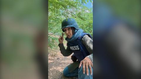 A reporter wearing a blue press flak jacket and helmet is crouching in some bushes. She is pointing upwards.