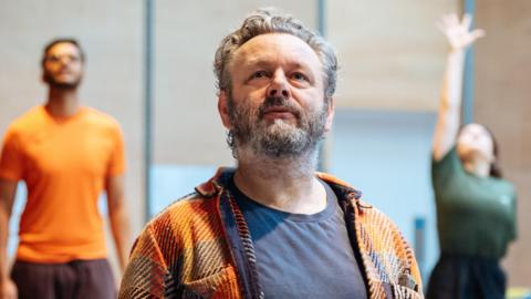 Michael Sheen in a checked shirt and T-shirt in the foreground, with other actors at the back of the rehearsal room