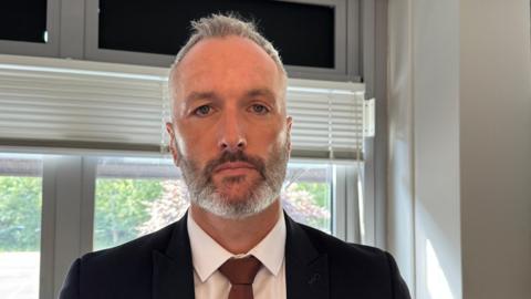 Det Con Lee Harshey-Jones. He looks at the camera with a neutral expression, and wears a white shirt, black suit jacket and bronze tie. He has short grey hair and short stubble.