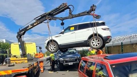 A white car which is being lifted by a truck. There are men in hi-vis standing around and a red car and a black car nearby.